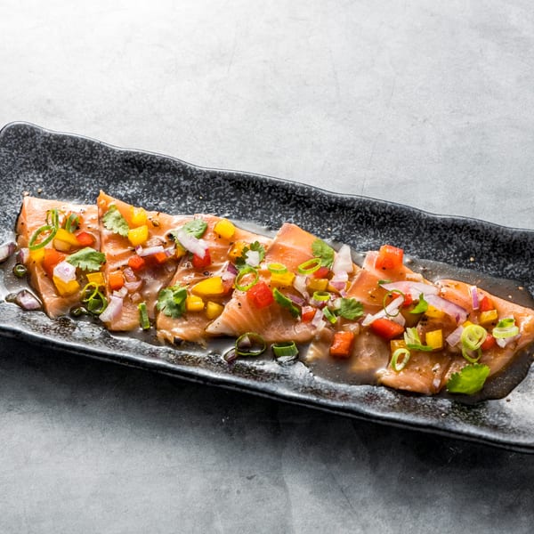 Salmon sashimi with colorful diced peppers, onions, and cilantro on a dark rectangular plate.