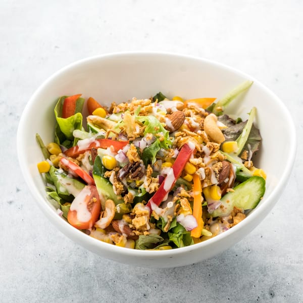 A vibrant salad with lettuce, bell peppers, corn, cucumber, nuts, and dressing in a white bowl.