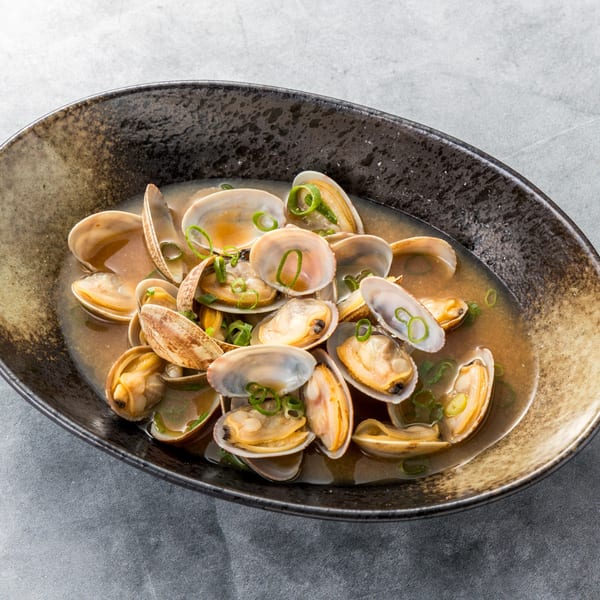 Clams in a savory broth with sliced green onions, served in a dark, textured bowl.
