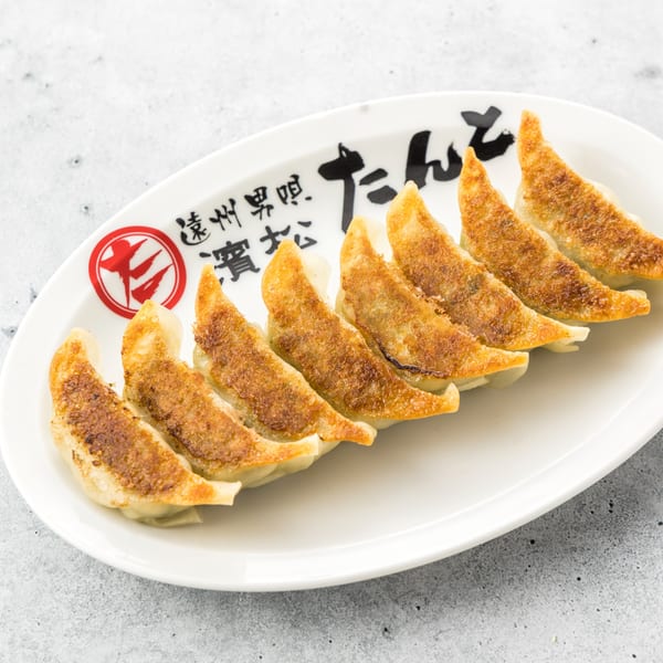 Seven golden-brown Tanto Gyoza dumplings arranged in a row on a white oval plate with Japanese writing.